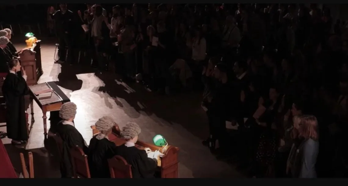 A darkened hall set as a courtroom, with barristers in wigs at the tables and a large audience behind