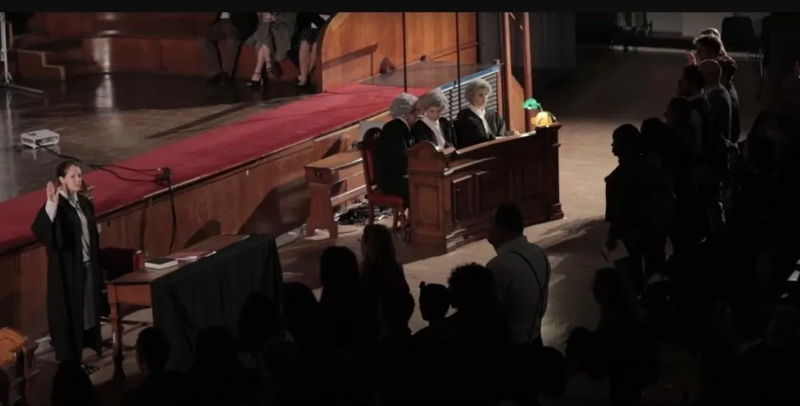 A witness with a raised hand in the courtroom set, watched by a seated audience