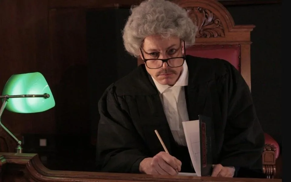 An actor as the judge, in wig and glasses, writing at the bench beside a green desk lamp