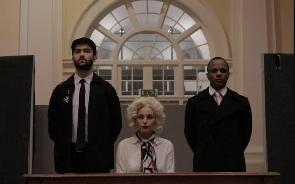 An actor with a blonde 1950s hairstyle in the dock, standing between two men in dark coats