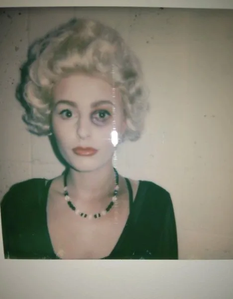 A portrait photograph of a woman with blonde 1950s curls, a bruised eye and a green top