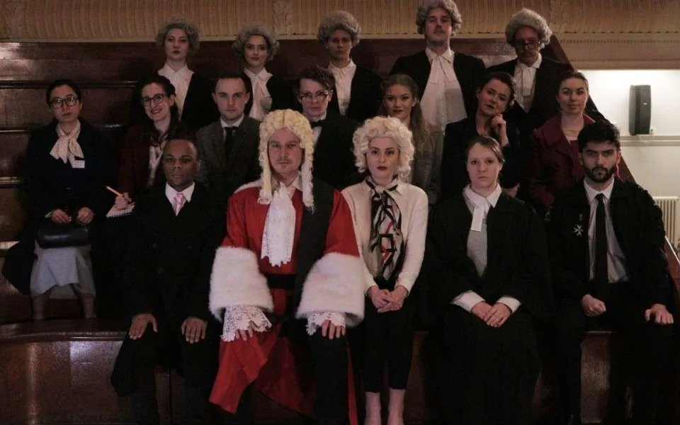 Performers in 1950s costume, barristers' wigs and a judge's red robe, posed on tiered benches