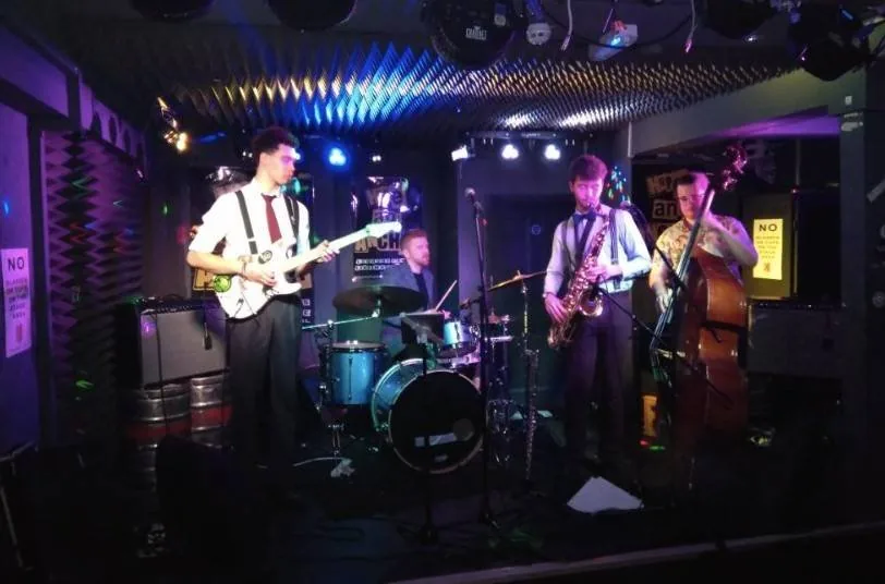 A band with guitar, drums, saxophone and double bass playing on a small stage