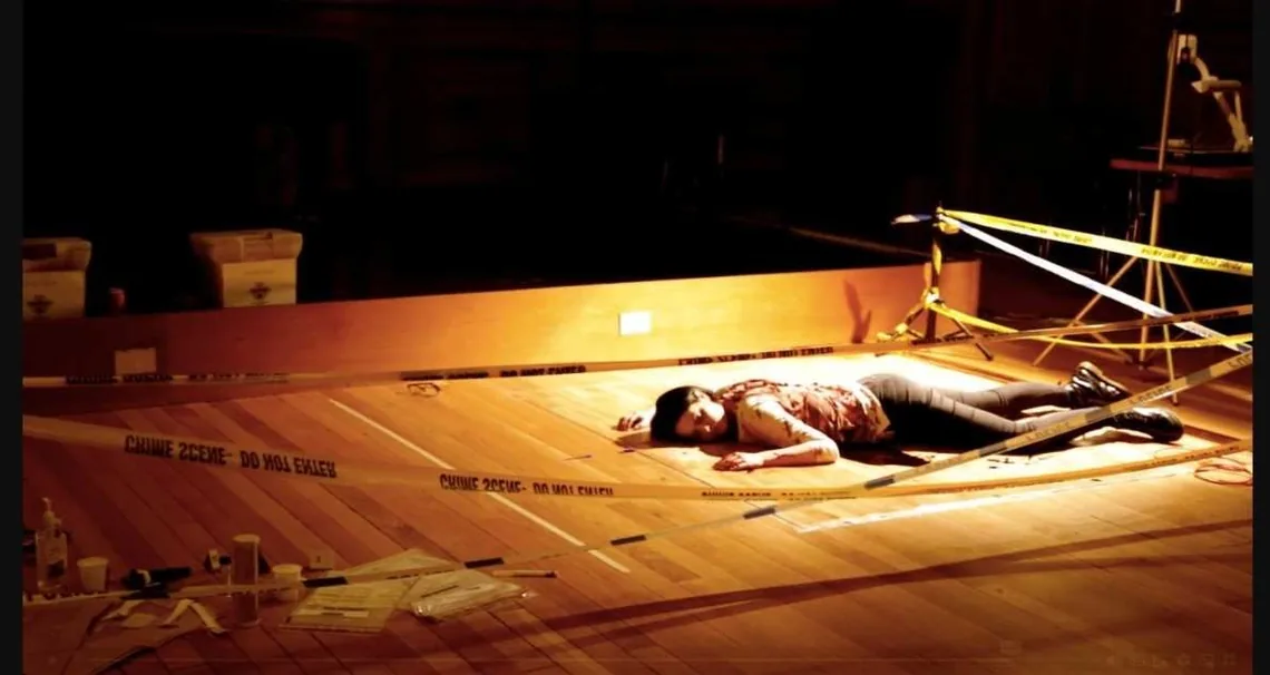 A staged crime scene on a stage: police tape, evidence boxes and a performer lying on the floor