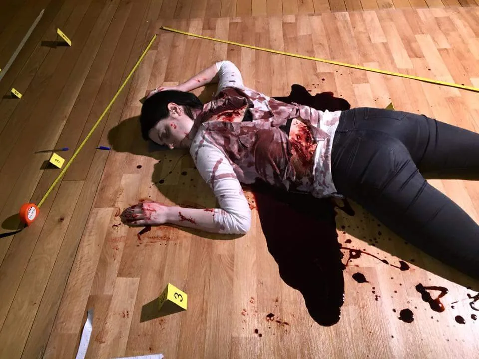 A performer lies on a wooden floor as the victim, surrounded by fake blood, evidence markers and a measuring tape