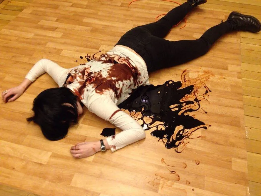 A performer lies face down on a wooden floor in a white top stained with fake blood