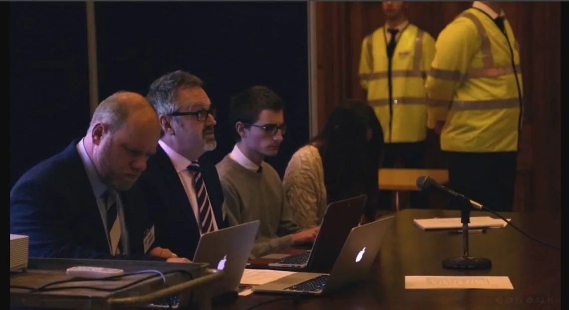 Four people in suits at a table with laptops and a microphone, two people in high-visibility jackets standing behind them