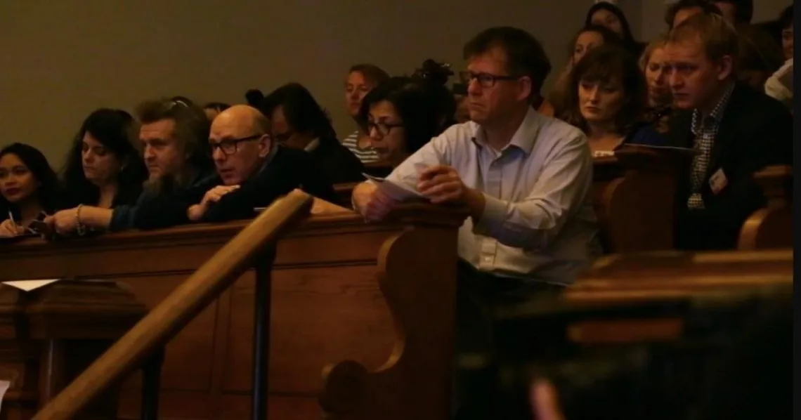 Audience members seated in wooden pews, watching closely