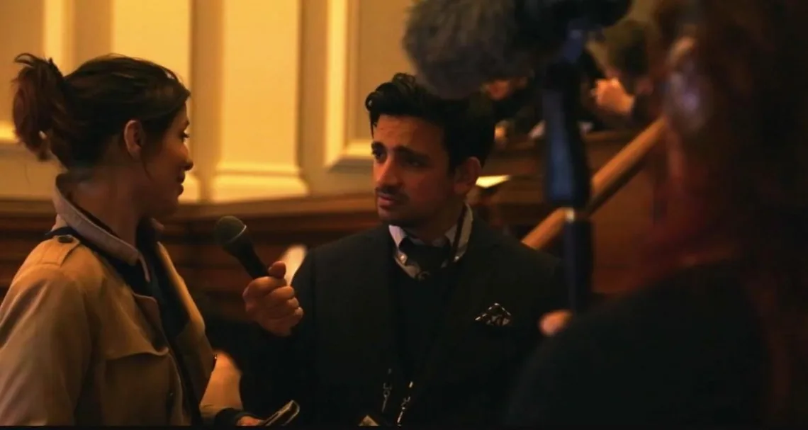 A man holding a microphone talks to a woman in a trench coat, a boom microphone overhead