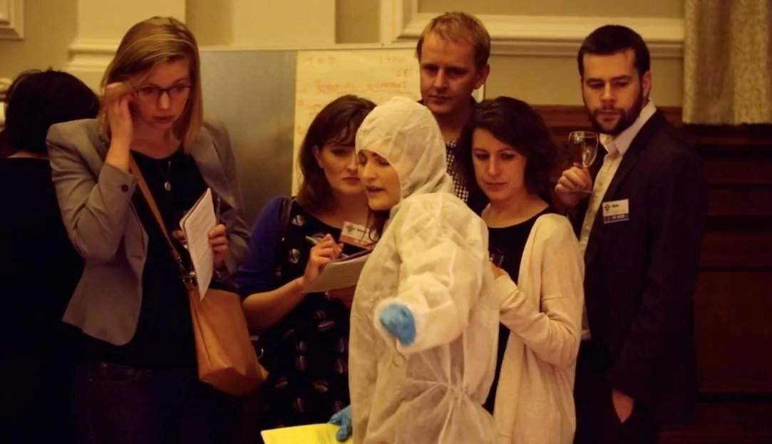 Audience members holding notes gather around a person in a white forensic suit