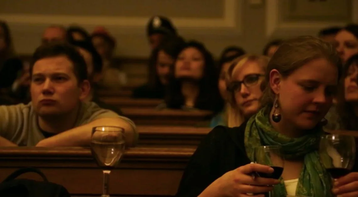 Audience members seated in wooden pews, some holding glasses of wine