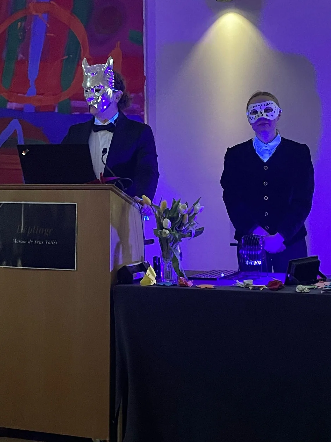 The masked auctioneer at the lectern and a masked assistant beside a table of lots
