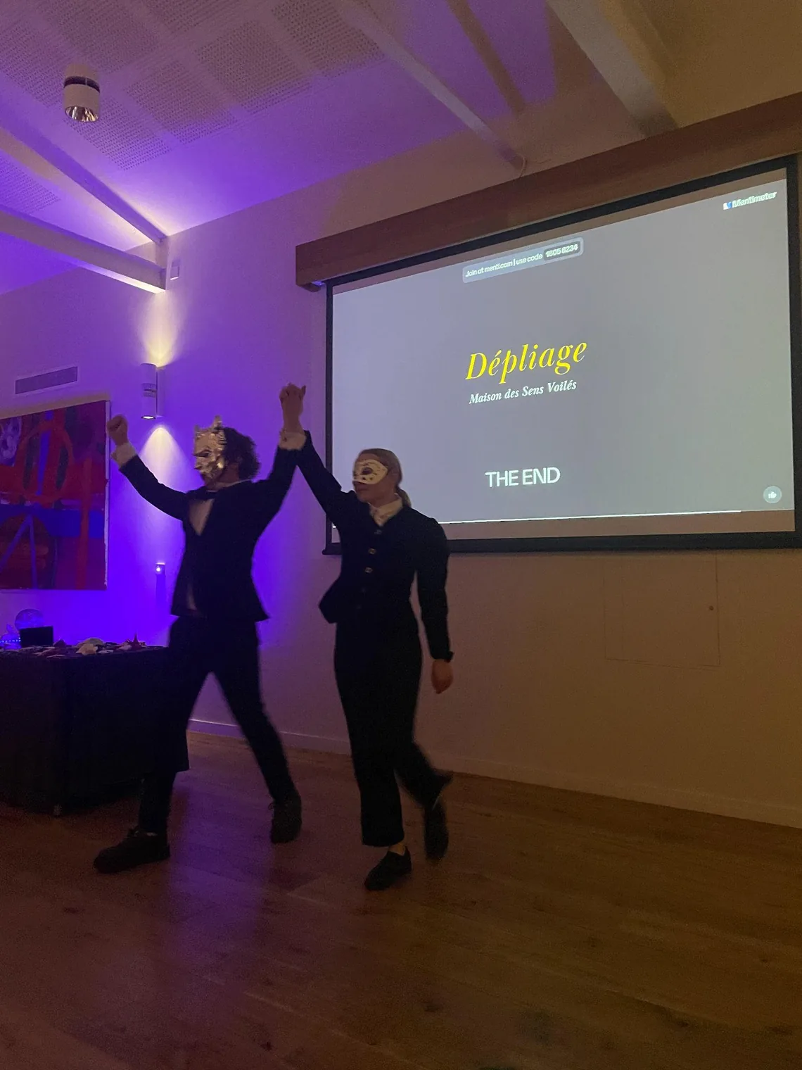 Two performers raise their arms in front of a screen reading 'Dépliage' and 'The end'