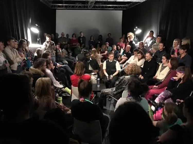 An audience seated close together in a room with black curtains and studio lamps