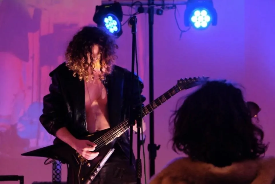 A long-haired guitarist in a leather jacket playing under blue stage lights