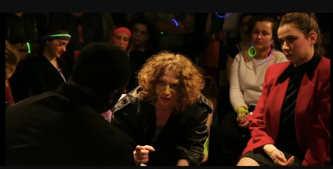 Performers in 1980s costume confront each other among an audience wearing glow sticks