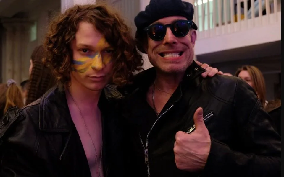 Two men in leather jackets pose for the camera, one with face paint, the other giving a thumbs up