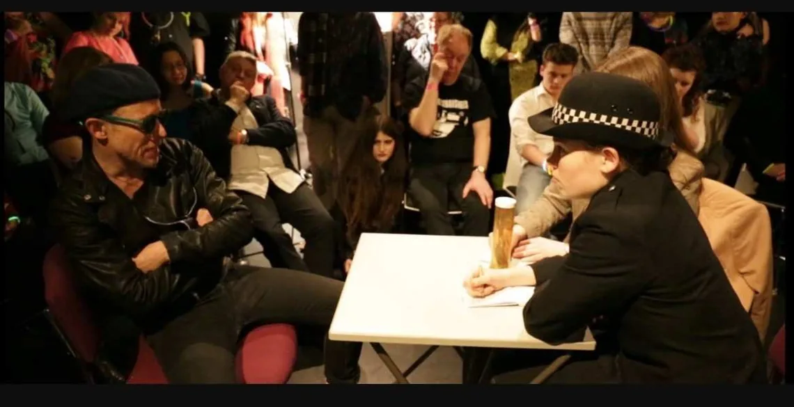A man in sunglasses and a leather jacket sits across a table from an actor in police uniform, the audience around them