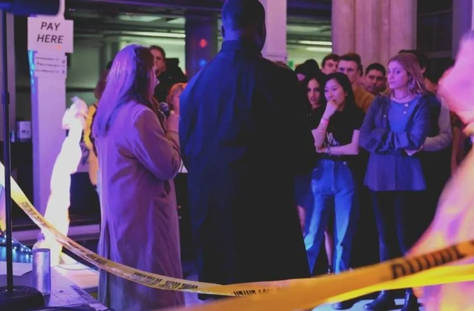 A woman speaks into a microphone behind yellow tape while a standing audience watches