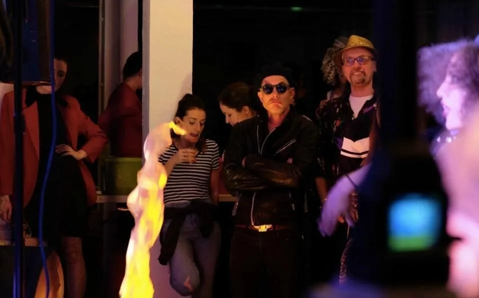 Audience members standing beside a pillar, a flame in the foreground